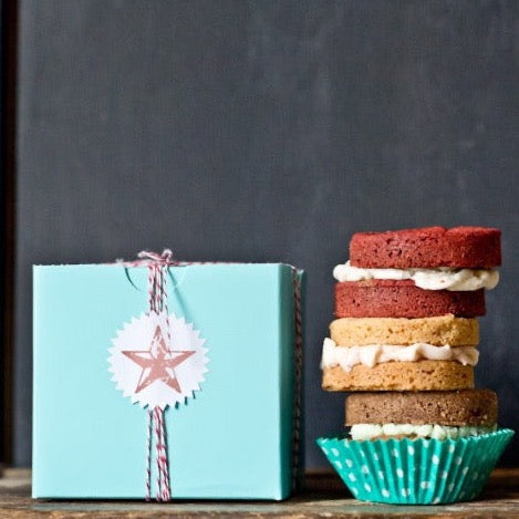 turquoise square 4x4 cupcake box wrapped with red and white striped bakers twine and a white starburst sticker label next to a stack of whoopie pies and a glass of milk