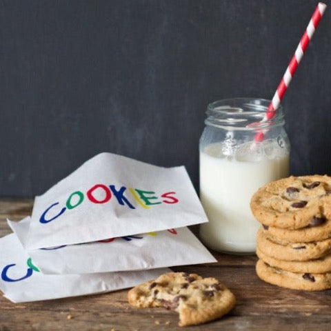 white glassine bags with rainbow cookies printed on the front next to a stack of chocolate chip cookies and a bottle of milk