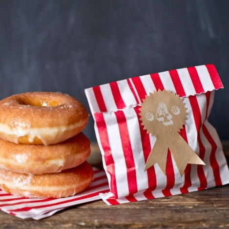 tan kraft sticker labels in an award ribbon shape next to red and white striped paper bags and a stack of glazed donuts