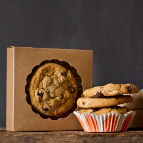 individual tan kraft cardboard cookie boxes with circle scallop cutout window next to a stack of chocolate chip cookies in an orange tulip cupcake liner