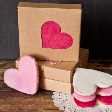 individual tan kraft cardboard cookie boxes with heart cutout window next to a stack of heart frosted sugar cookies