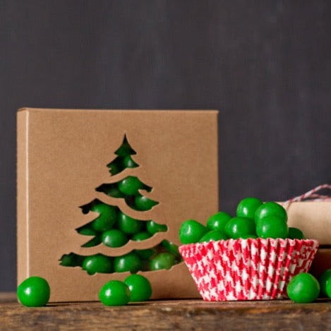 individual tan kraft cardboard cookie boxes with Christmas tree cutout window next to red and white houndstooth cupcake liner filled with candies