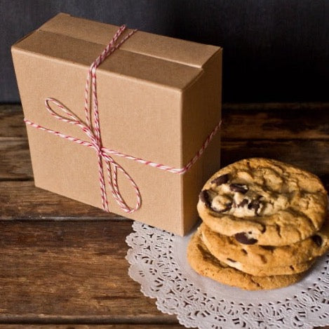 individual tan kraft cardboard cookie box wrapped with red and white striped bakers twine and next to a stack of chocolate chip cookies
