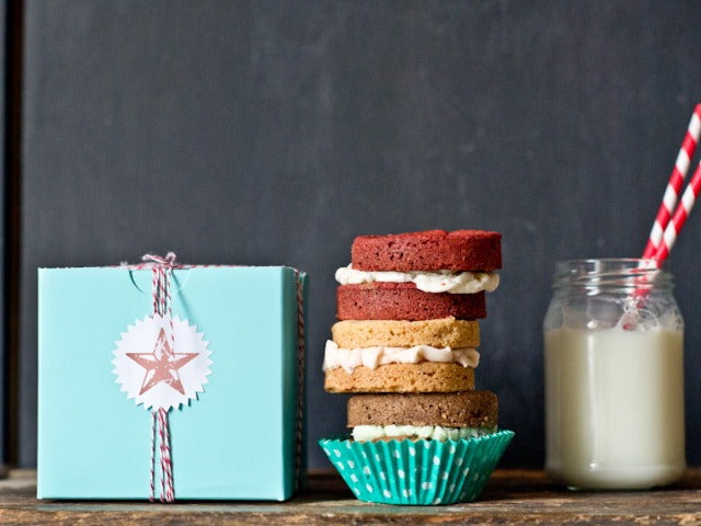 turquoise square 4x4 cupcake box wrapped with red and white striped bakers twine and a white starburst sticker label next to a stack of whoopie pies and a glass of milk